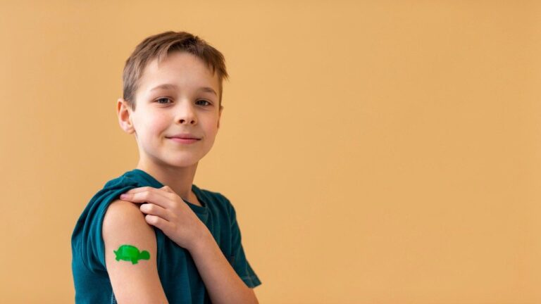 A smiling boy holds up his T-shirt sleeve to show a green turtle-shaped bandage on his upper arm.