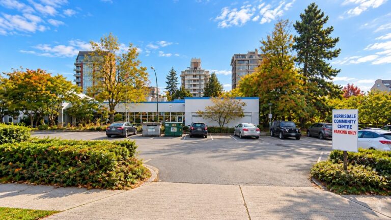 A parking lot in front of a low white building, with a sign reading "Kerrisdale Community Centre Parking Only", autumn trees and a blue sky.